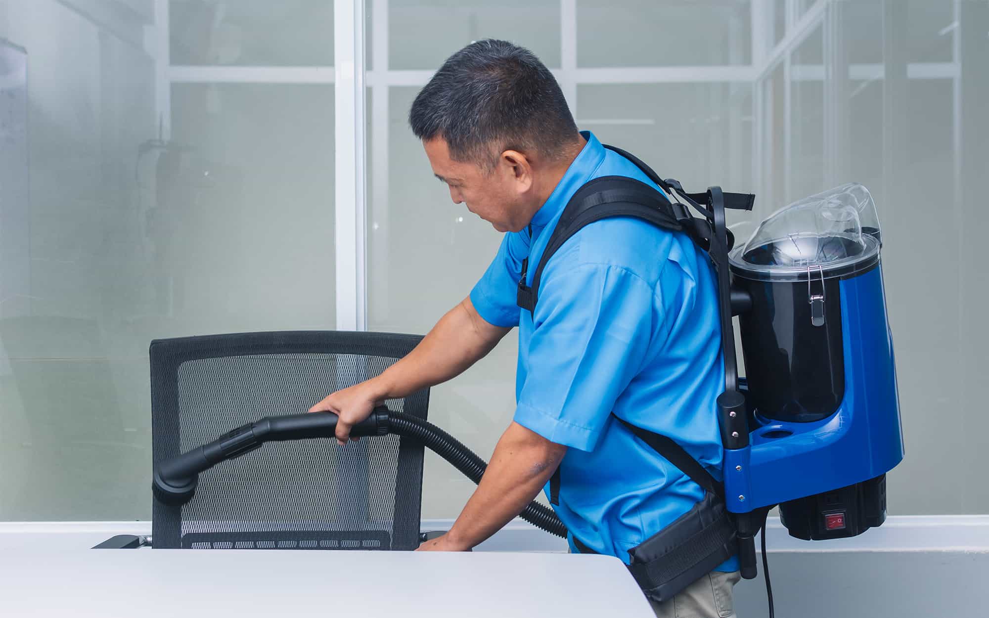 A Professional Janitor In A Blue Uniform Using A Commercial Backpack Vacuum To Clean A Modern Office Seat